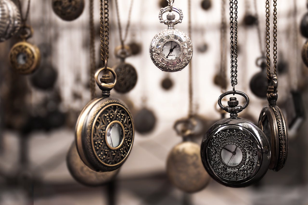 heros-img Close-up of antique pocket watches with intricate designs hanging in a blurred vintage setting.